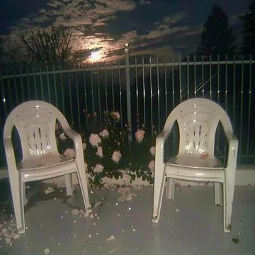 Two white chairs and flowers beside a moonlit fence.